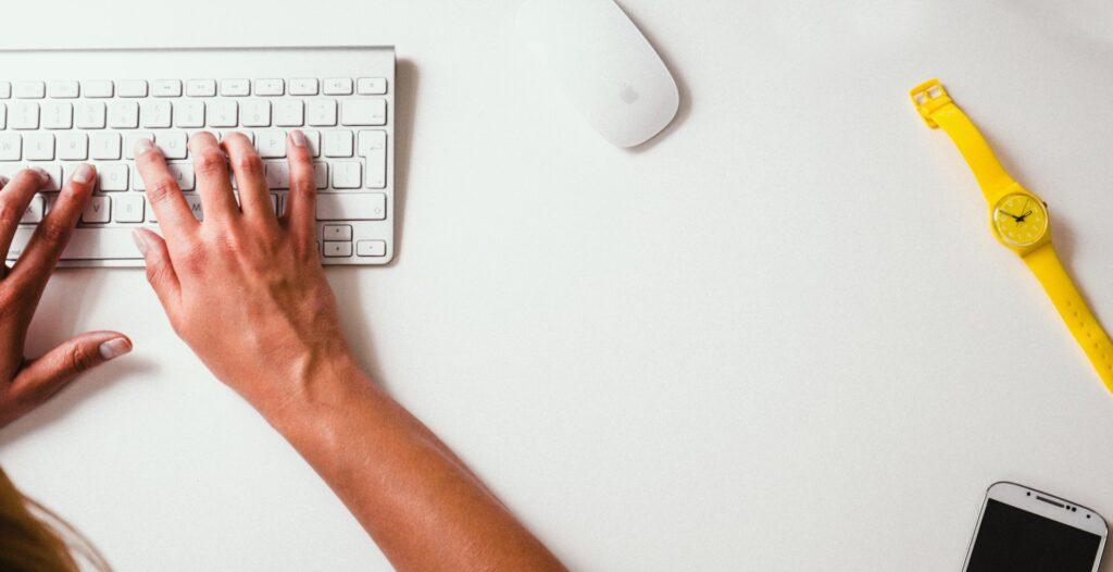 Nahaufnahme von Händen auf einer Tastatur, die an einem Webprojekt arbeiten, gelbe Uhr und Smartphone auf dem Tisch, Internetagentur Düsseldorf