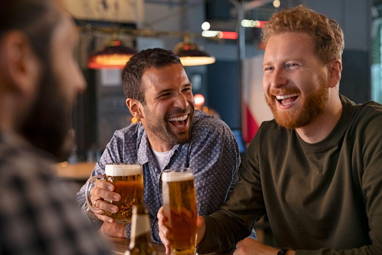 Drei Männer genießen ein Glas Tripel Karmeliet in einer gemütlichen Bar, lachen und stoßen an – das perfekte Bier für gesellige Abende und echte Genießer.