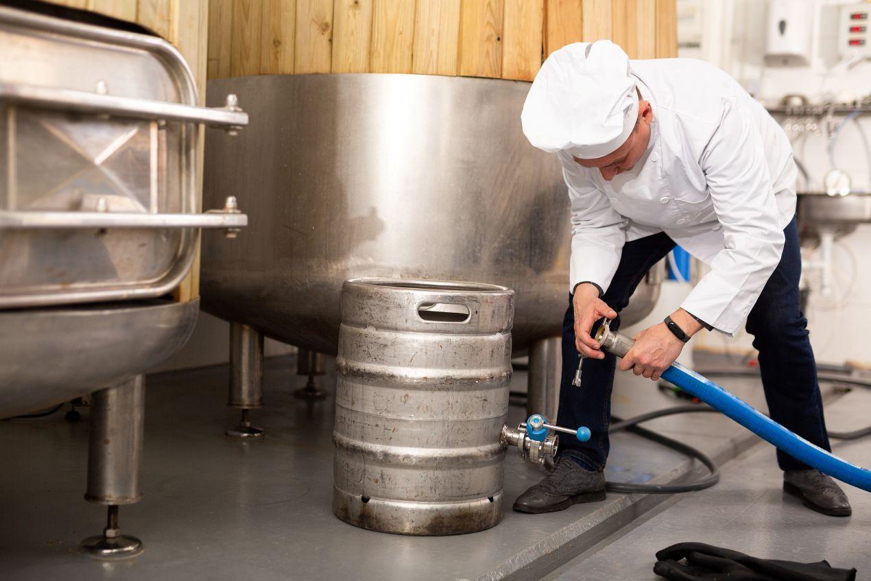 Ein Braumeister in weißer Uniform schließt ein Metallfass an, um frisches Tripel Karmeliet aus einem Gärtank abzufüllen – Abfüllprozess in einer traditionellen belgischen Brauerei.