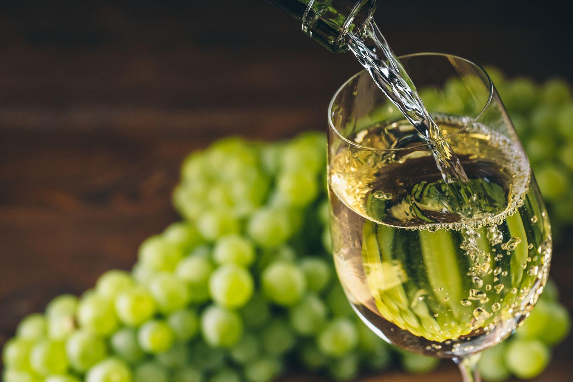 Klarer Weisswein fliesst in ein Glas mit hellgruenen Trauben im Hintergrund